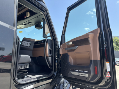 Truck cab interior view from open driver door. Door panel shows beige/brown trim with 'MACK' branding on door sill. Dashboard area visible with steering wheel and controls.