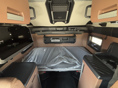 Truck sleeper cab interior with brown leatherette bunks and overhead storage, center bed covered in plastic, control panel and vents visible.