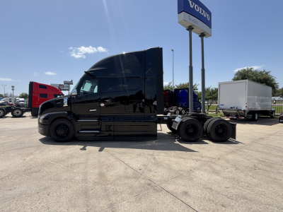 Truck tractor in black, side view showing sleeper cab and rear axles with dual wheels; dealership lot in background with tall Volvo sign.