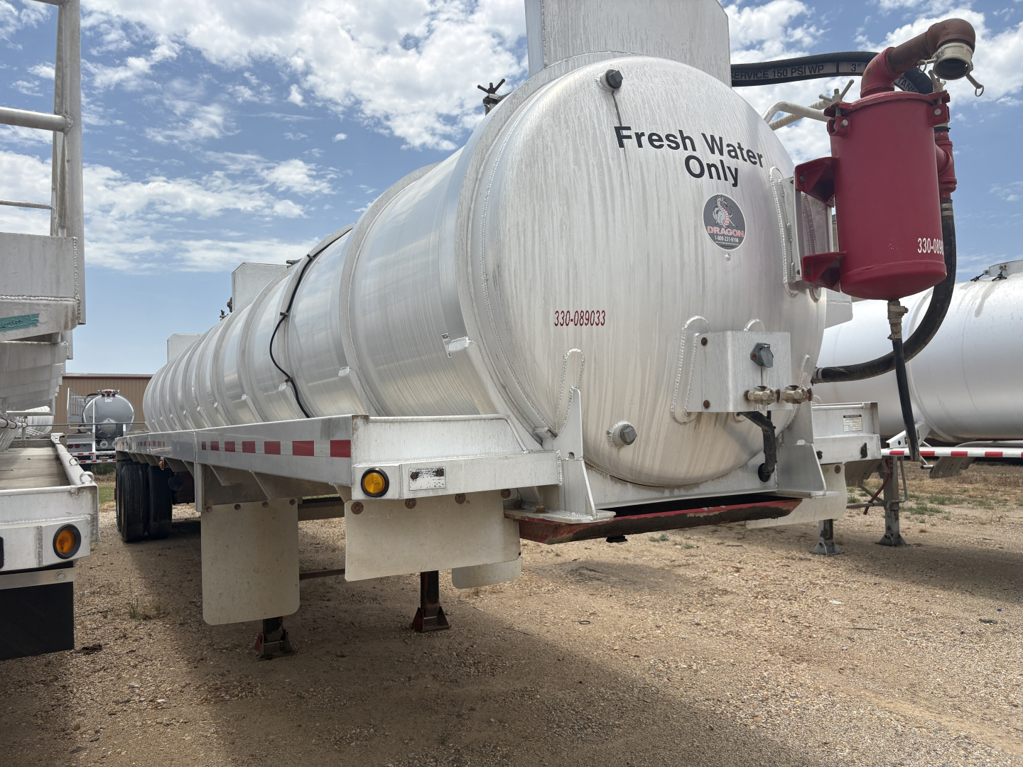 Tank trailer showing a cylindrical aluminum vacuum tank with text Fresh Water Only and a red pump on the rear, mounted on a greenish-white trailer with tires, dirt ground, cloudy sky.