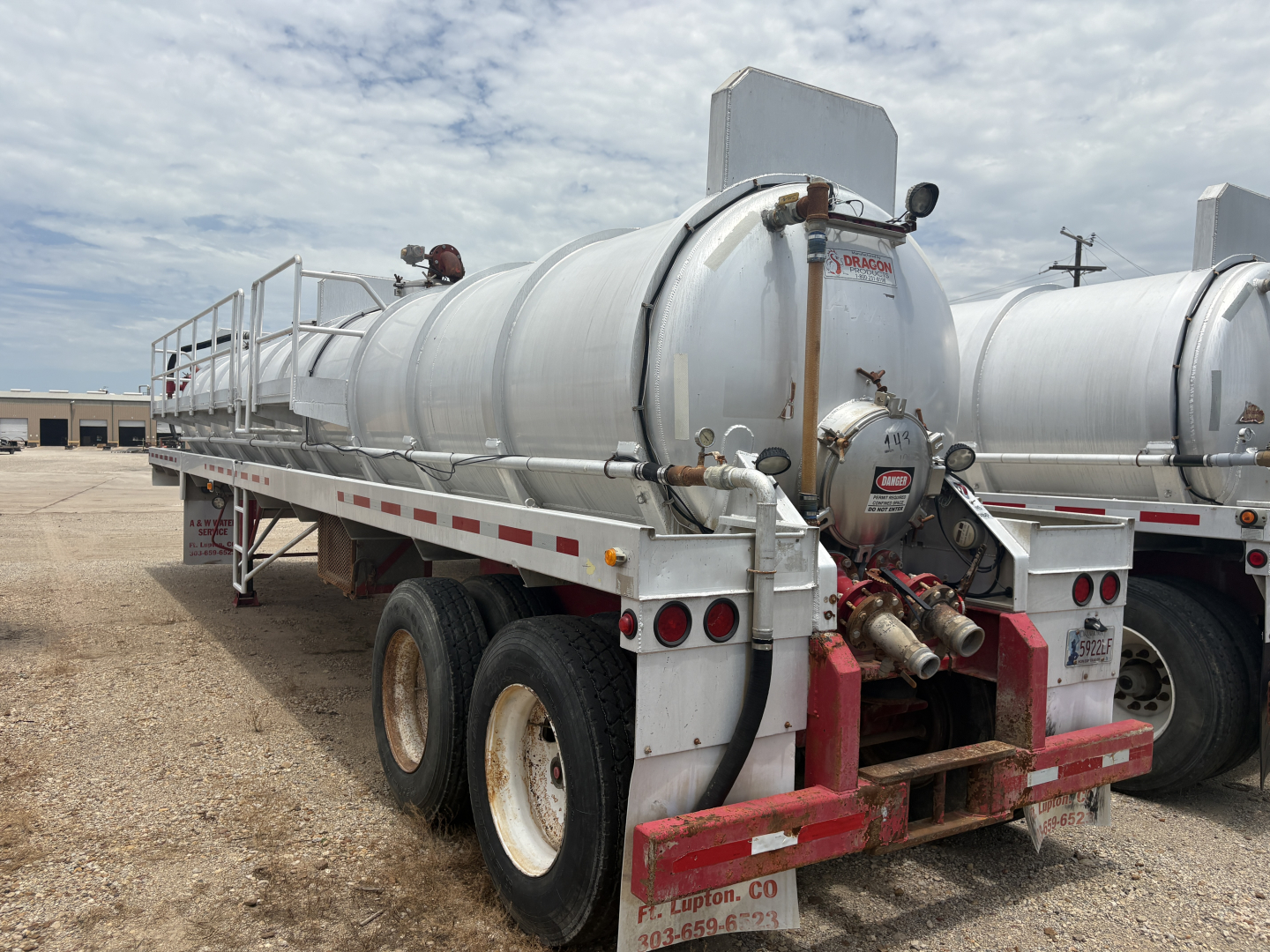 Vacuum trailer with a cylindrical stainless tank mounted on a heavy-duty dual-axle chassis, rear red bumper, piping and valves, and rear safety lights.