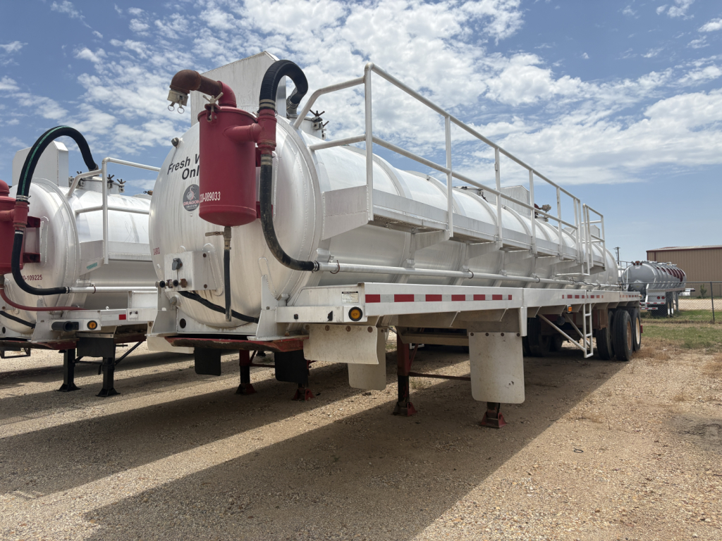 Vacuum trailer with large cylindrical tank and red fire extinguisher mounted at rear, metal railings along top, white exterior, parked on dirt lot.