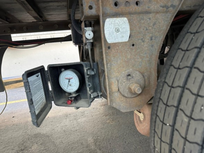 Trailer underside showing a pressure gauge in a protective case mounted near the wheel; rusted frame and hitch area.