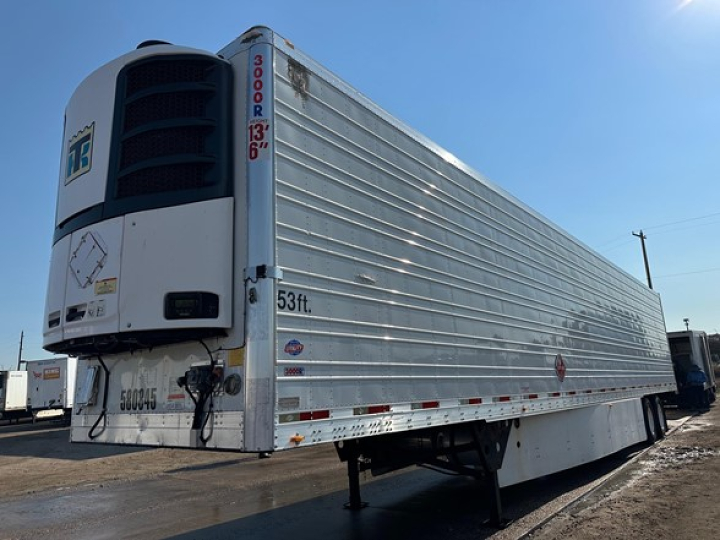 Trailer side view showing a large refrigerated trailer with a white Thermo King unit at the front, ribbed aluminum sides, and adjustable landing gear extended.