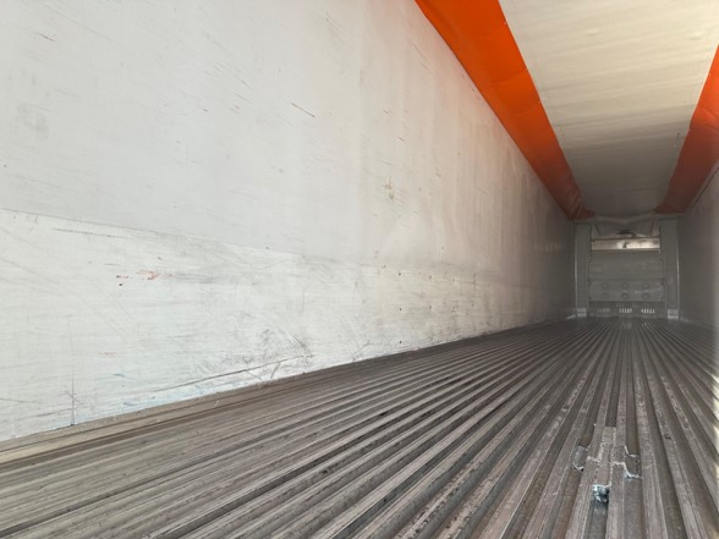 Trailer interior showing empty refrigerated trailer with corrugated floor and white walls, orange ceiling beams, and rear cargo door.