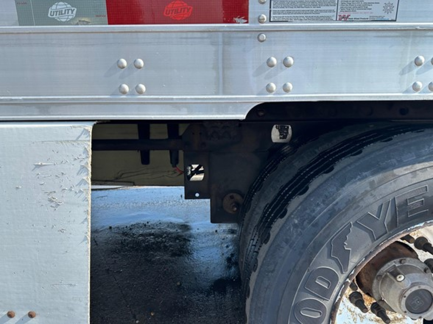 Trailer rear corner showing stickered utility branding, metal frame, and dual-axle wheel with tire; partial view of undercarriage and reflective surfaces.