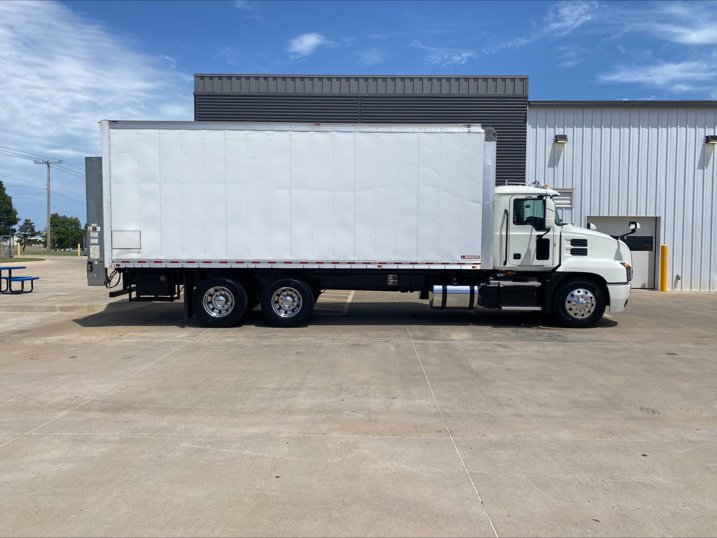 Truck side view of a white 2019 Mack AN64 box truck, cab with sleeper, long box body, parked on concrete in front of a building.