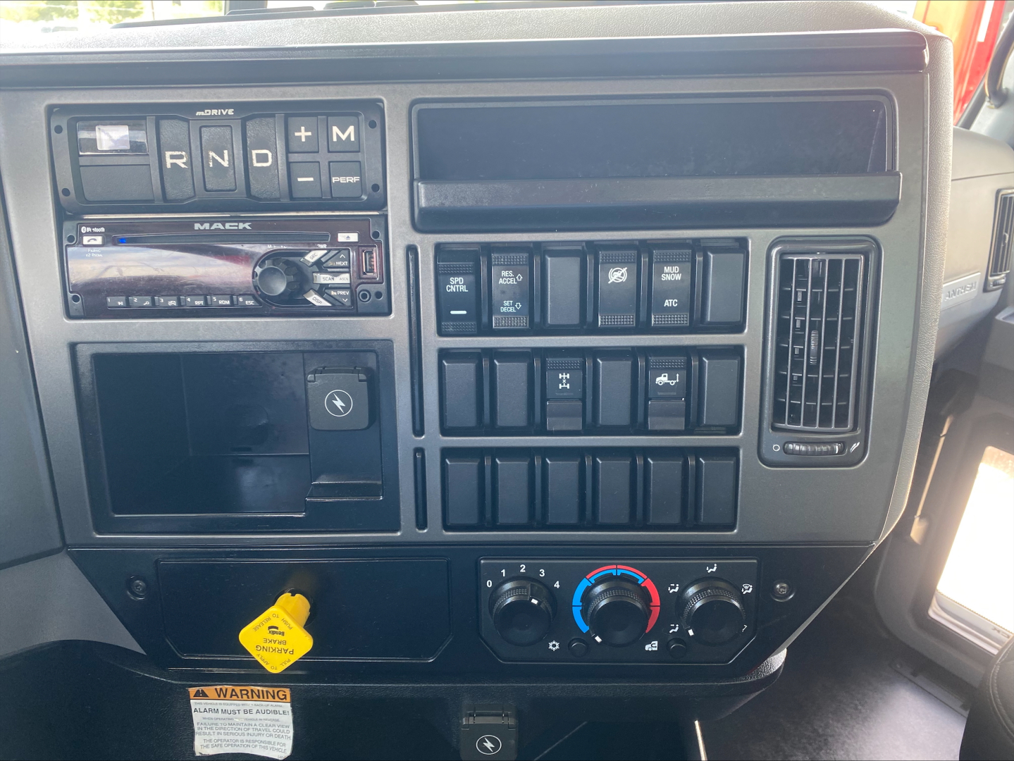 Dashboard of a 2019 Mack AN64 truck showing gear selector, various control switches, climate controls, and a yellow electrical plug socket.