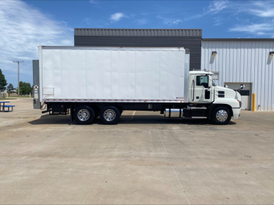 Truck side view of a white 2019 Mack AN64 box truck, cab with sleeper, long box body, parked on concrete in front of a building.