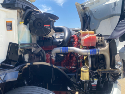 Engine bay of a 2019 Mack AN64 in-service showing red engine, intercooler piping, coolant reservoir, and yellow oil filter with blue hose clamps.