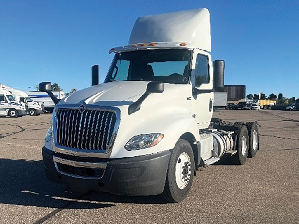 White 2019 International LT625 daycab truck front-left view; cab roof fairing; large chrome grille; black front bumper; side mirrors; dual rear axles; empty fifth wheel area; background shows other trucks in lot.