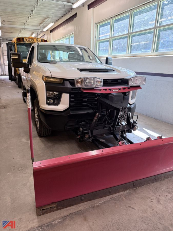 Front view of a white 2022 Chevrolet Silverado 3500HD pickup with a pink snow plow attached; plow mounting hardware visible; vehicle inside a service bay with a school bus in background; no visible text readable on the vehicle except the pl