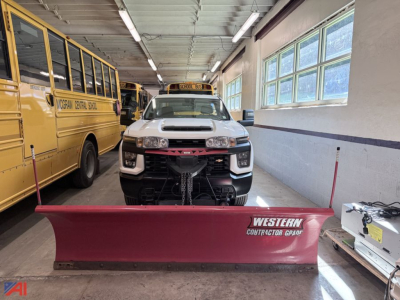 Chevrolet Silverado 3500HD pickup with plow attachment in a workshop showroom, front view, yellow school buses nearby, plow labeled Western Contractor Grade.