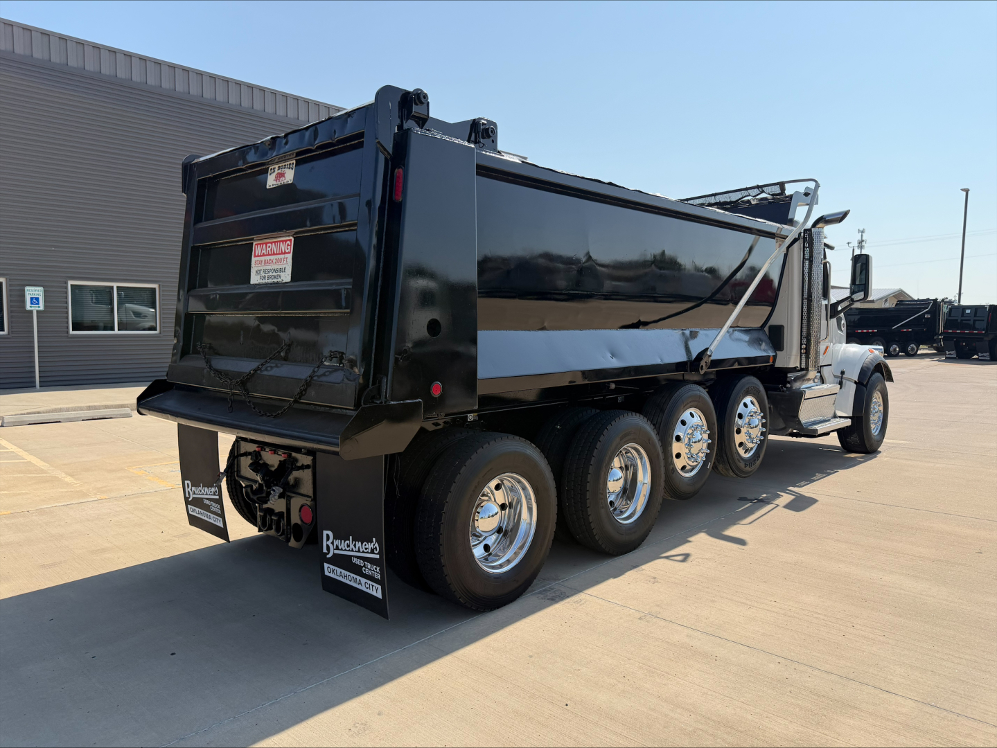 Peterbilt 567 dump truck shown from rear-three-quarter view with a black dump body, triple rear axles, and chrome wheels.