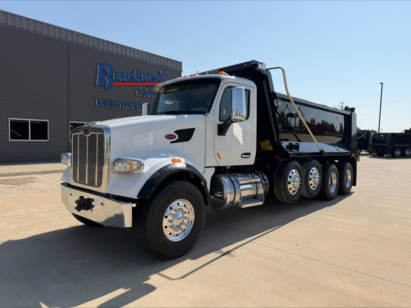 Peterbilt 567 dump truck in a dealership lot, white cab, black dump bed, multiple axles, chrome bumper and wheels, front grille with Peterbilt badge.