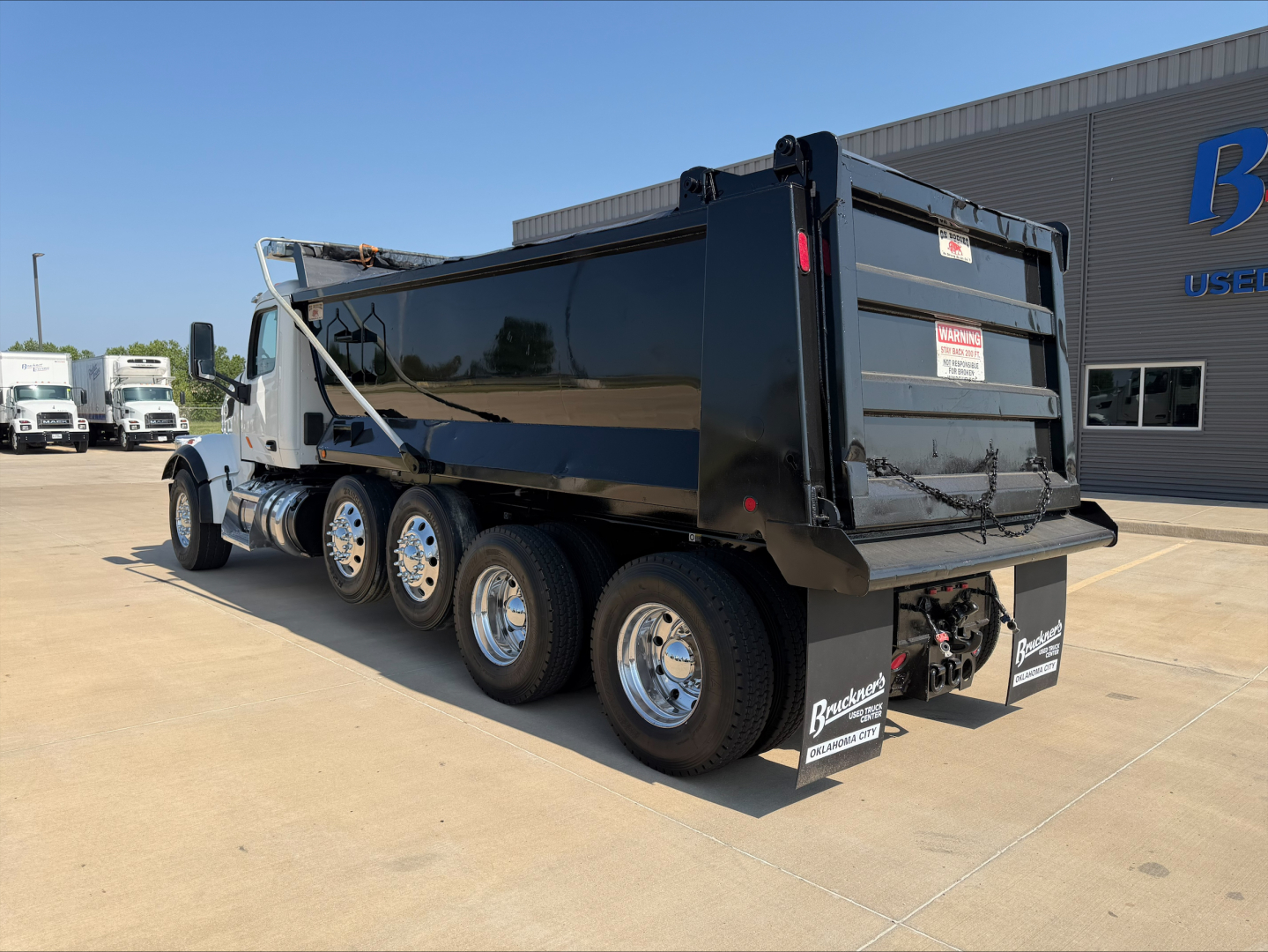 Peterbilt 567 dump truck in service showing side and rear with black dump bed, chrome wheels, rear bumper, and stickers on mud flaps.