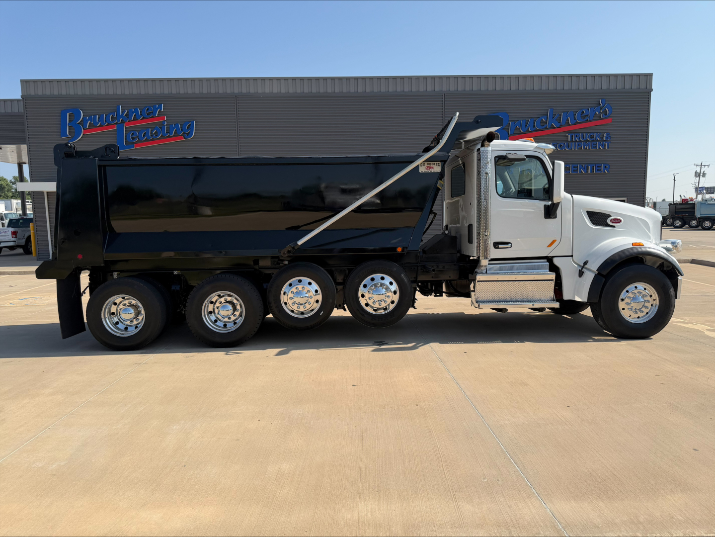 Peterbilt 567 dump truck side view showing black dump body, white cab, chrome wheels, and 'Bedrock' dealership signage in background.