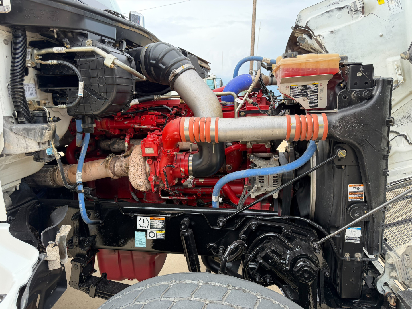Peterbilt 567 diesel engine bay with red engine, black radiator, blue hoses, and orange intake tube; visible battery and alternator; front wheel and chassis components.