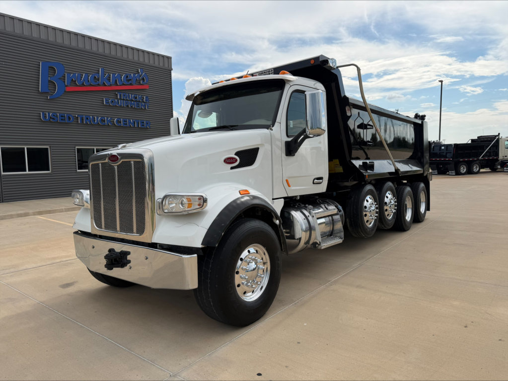 Peterbilt 567 dump truck in front of dealership with white cab, chrome bumper, and multiple axles; Bruckner’s Used Truck Center signage visible.