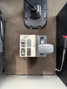 Panel with labeled switches and a knob on a carpeted truck interior wall, showing a ‘DE NOBLES’ panel and a toggle/rocker switches, gear selector nearby.