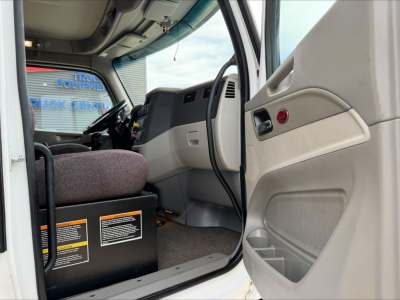 Peterbilt 567 truck interior cabin viewed from open driver door, dashboard and steering wheel visible, orange and black warning labels on lower dash near pedals.