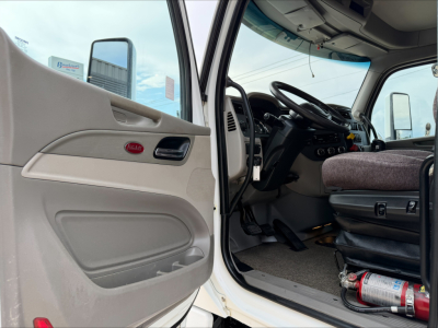 Peterbilt truck interior showing driver's seat and open door with panel, steering wheel, dashboard, and Pedal area; seating and blue-gray fabric visible.