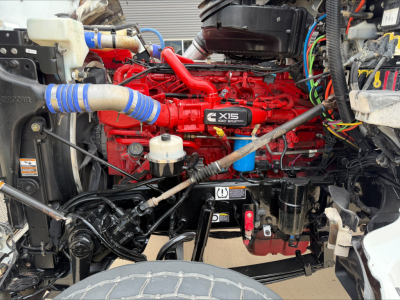 Peterbilt 567 engine bay with bright red Cummins X15 diesel engine, various hoses, wiring, and front suspension visible.