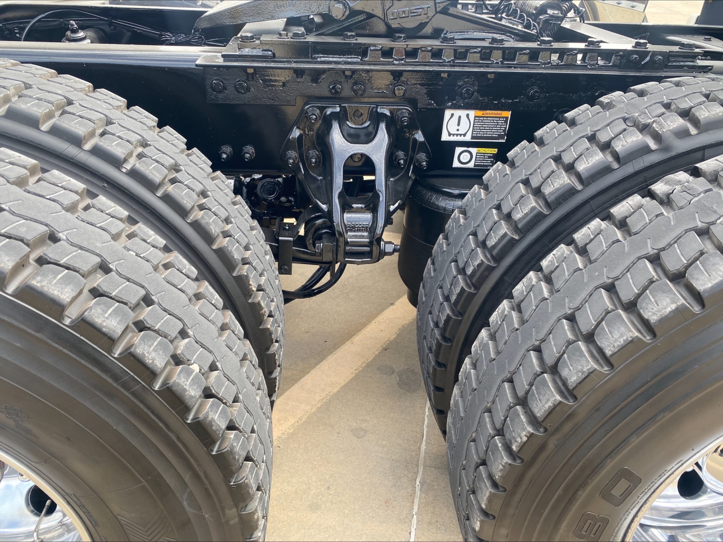 Truck chassis with dual rear axles and fifth wheel mounting point visible; close-up shows suspension and coupling assembly between the two rear wheels.
