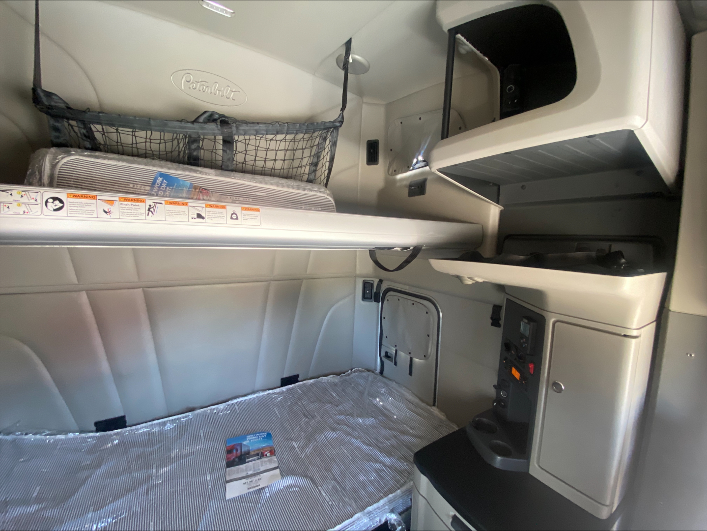 Interior view of a 2022 Peterbilt 579 sleeper cab showing the upper bunk and storage compartments with nets, controls, and a desk area.
