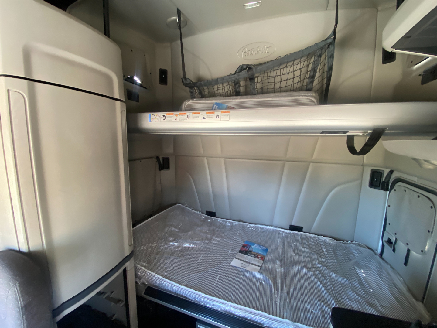 Peterbilt 579 cab interior sleeper bunk area with mattress and overhead storage shelf, side net, and ladder support visible.