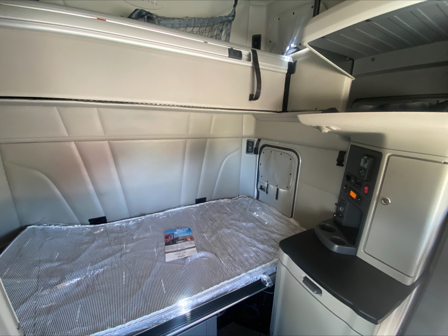 Interior sleeper cab of a Peterbilt 579 showing bunk bed with plastic wrap, overhead storage, and control panel on right; light gray panels and curved surfaces, compact workspace.