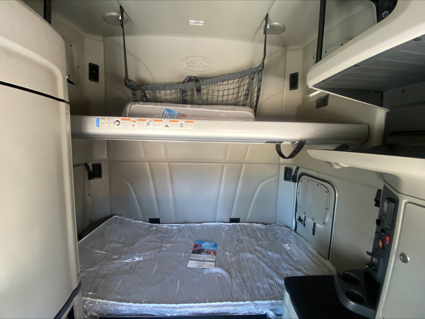 Cab interior of a 2022 Peterbilt 579 sleeper with cargo net, overhead storage, and a bed covered in plastic.