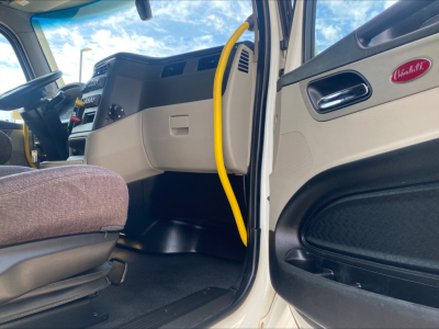 Peterbilt 579 interior cab area showing steering wheel, dash, beige door panel, yellow air hose, and driver seat. Close-up of entry with Peterbilt emblem on door panel.