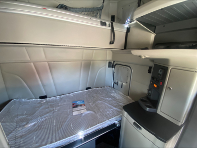 Interior sleeper cab of a Peterbilt 579 showing bunk bed with plastic wrap, overhead storage, and control panel on right; light gray panels and curved surfaces, compact workspace.