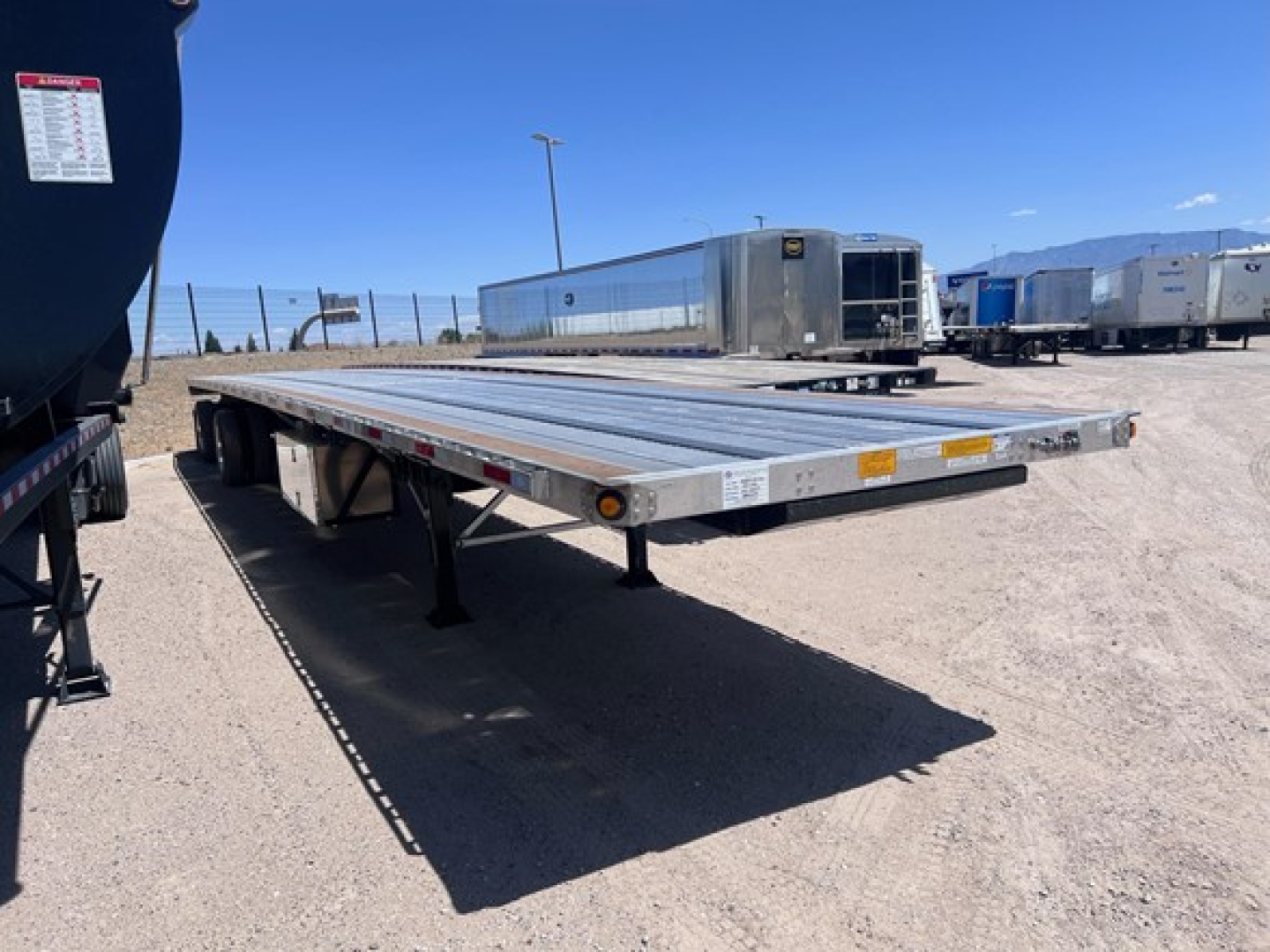 Flatbed trailer with a long aluminum deck viewed from side; other trailers and a refrigerated trailer in background; clear blue sky.