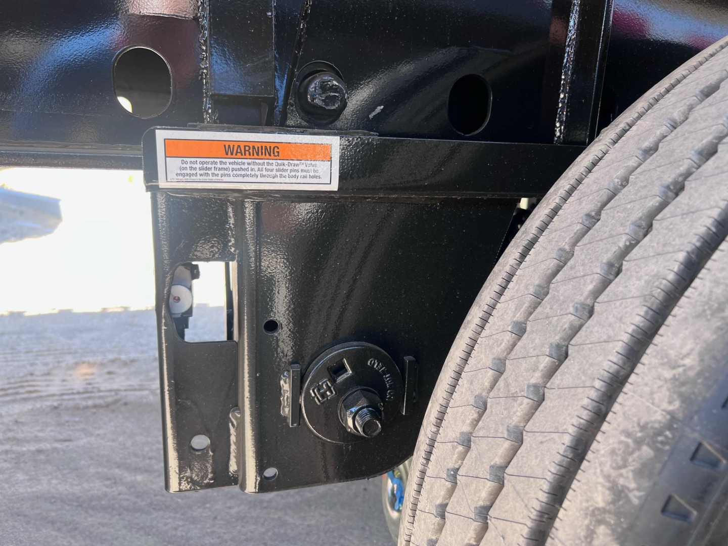 Flatbed trailer hitch area showing a circular PTO/bolts with label and warning sticker on black metal frame near a tire.