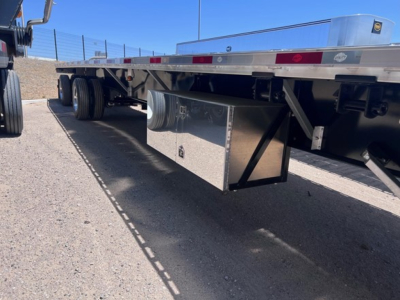 Flatbed trailer undercarriage showing toolbox/storage box and dual rear wheels, aluminum side rails, and reflective tape along frame.
