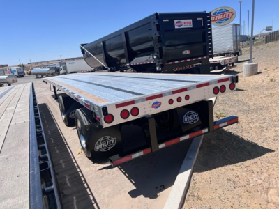 Utility flatbed trailer with aluminum deck and rear ramp, back view showing taillights, spare tire covers, and stickered rear bumper.