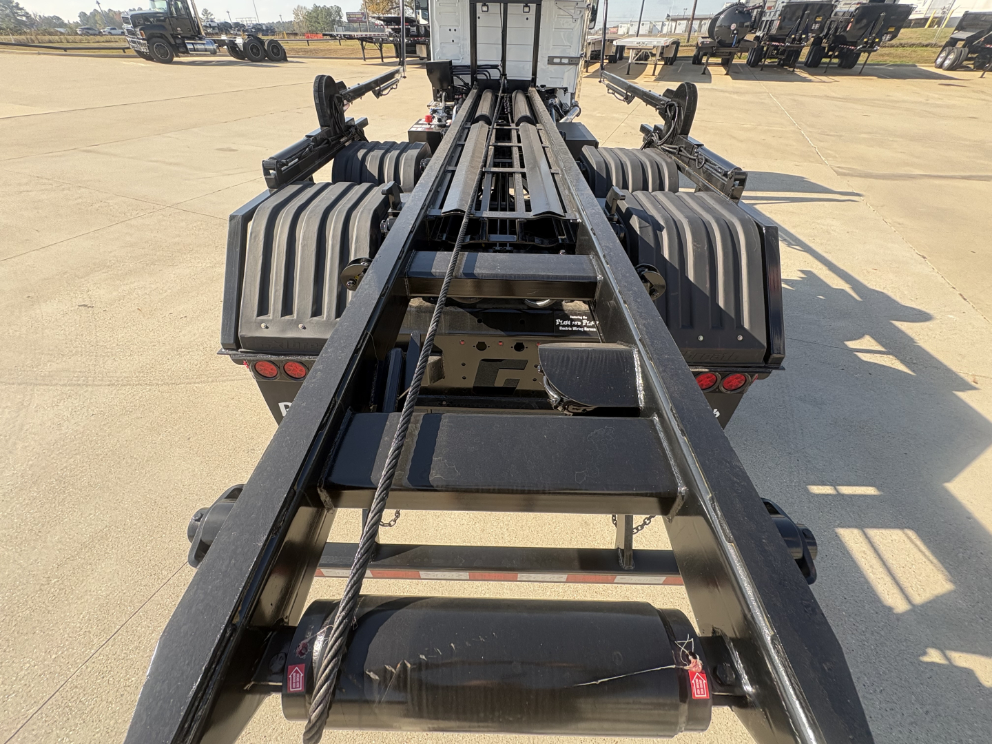 Truck chassis/rail frame of a Volvo VHD64B300 roll-off truck, with heavy steel rails and winch hardware visible along the center, crew cab rear in background.