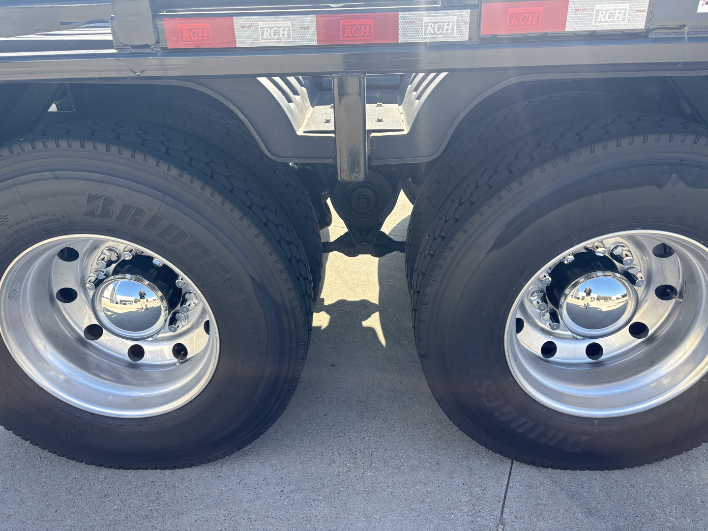 Dual rear wheels of a Volvo VHD64B300 heavy truck showing chrome hubcaps and dual tires, with trailer frame and reflectors above.