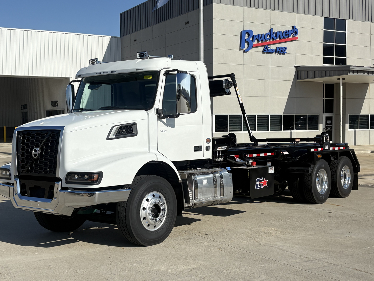 Volvo VHD64B300 roll-off truck in front of a dealership, white cab, chassis with multiple axles and hoist equipment visible.