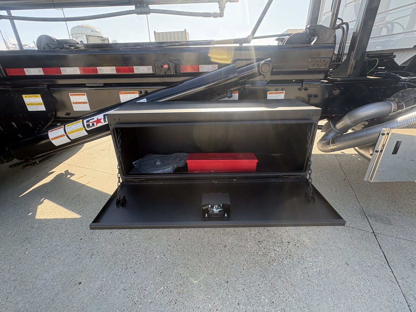 Open storage box on a Volvo VHD64B300 roll-off truck showing interior with red tool case and items. Side rails, hydraulic arm, and various stickers visible.