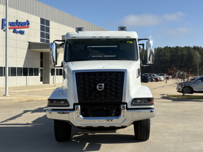 Front view of a white 2026 Volvo VHD64B300 truck in a dealership lot. Clear grille, large chrome bumper, Volvo emblem, side mirrors, and dealership building in background.