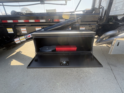 Open storage box on a Volvo VHD64B300 roll-off truck showing interior with red tool case and items. Side rails, hydraulic arm, and various stickers visible.