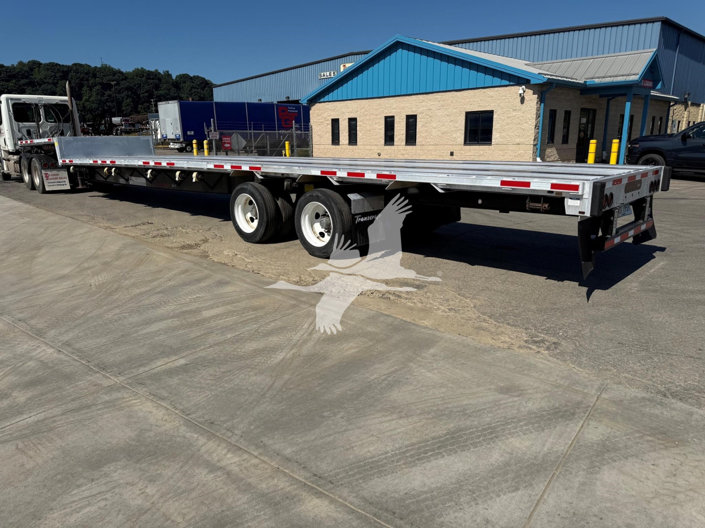 Trailer showing a long flatbed with dual rear axles, white and red safety stripes, parked at a facility.