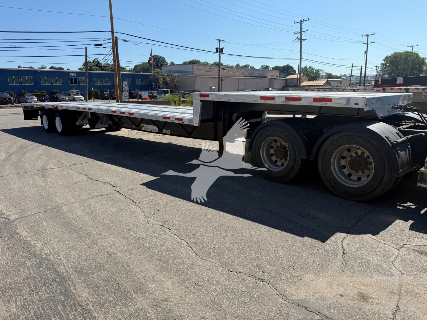 Trailer side view showing Transcraft D-Eagle II plate with long flatbed and dual rear axles. Visible labels include branding on the trailer and DOT-style markings along the rail.