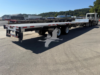 Flatbed trailer in a truck yard showing a long aluminum deck with rear bumper, taillights, and tandem axles under a tractor, 2020 Transcraft D-Eagle II branding on listing.