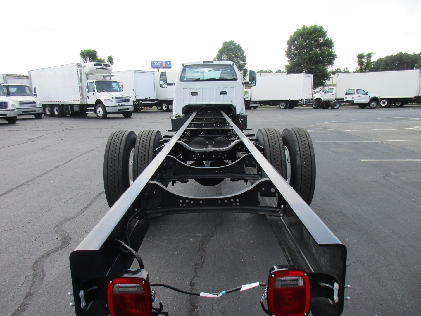 Front view of a Ford F650 SD cab and chassis frame showing chassis rails and dual rear axles, with a white cab in the distance, plenty of trucks in a lot.