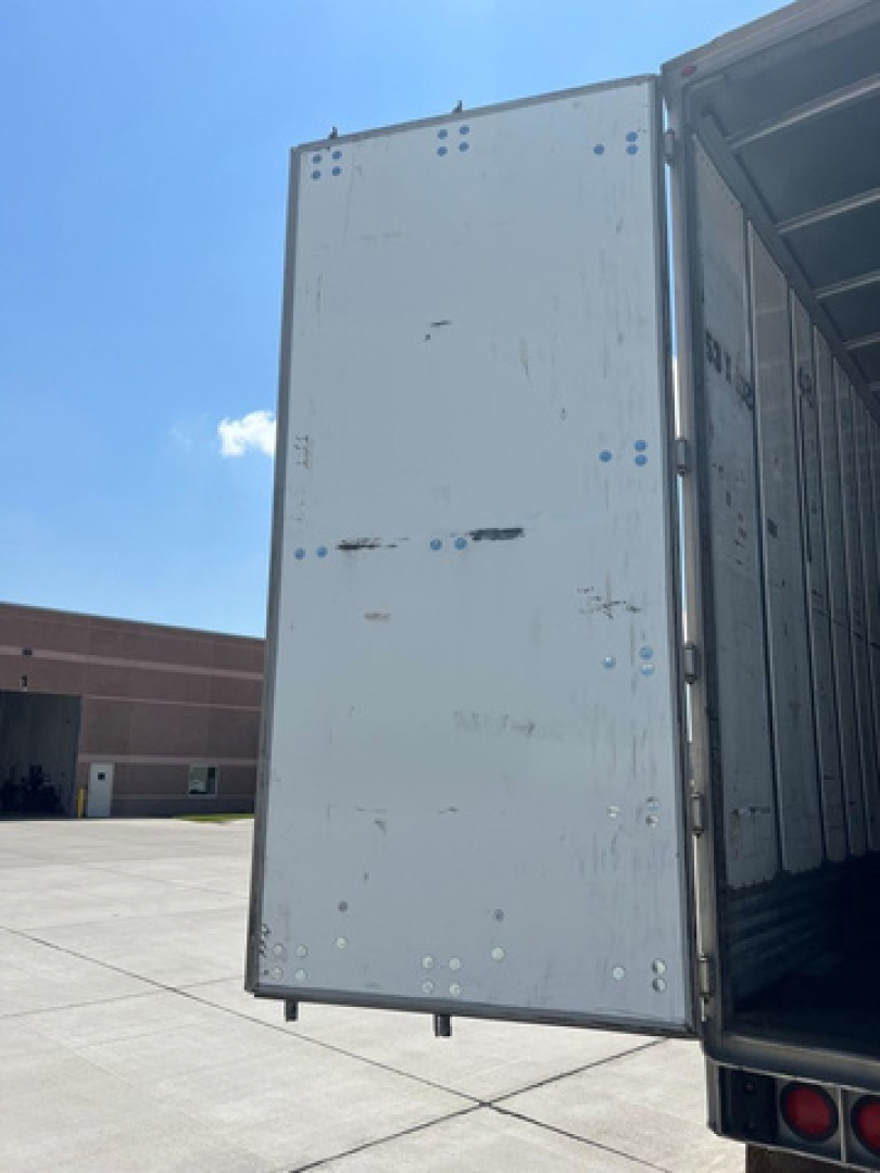 Trailer van rear interior showing back door and metal interior panel with rivets, light gray exterior, blue sky.