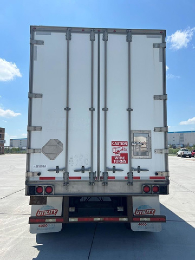 Rear view of a white utility trailer van showing double doors with metal latches, CAUTION sign, lights, and Utility brand mud flaps; license-free concrete lot.